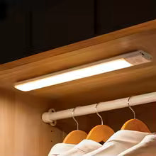 Under-cabinet light fixture illuminating a closet with hanging clothes.