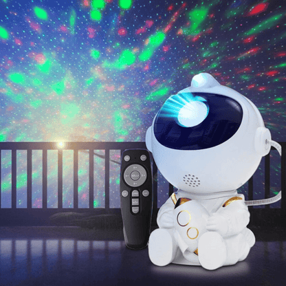 A white astronaut-shaped robot projector sits on a dark floor next to a black remote control, with colorful star and nebula light projections visible in the background.