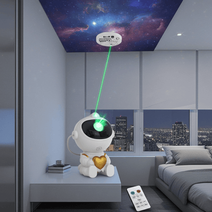 A small white robot-shaped projector sits on a bedside table in a modern bedroom, projecting a green laser light onto a galaxy-patterned ceiling. The scene includes a remote control on the floor, a bed, and city windows in the background.